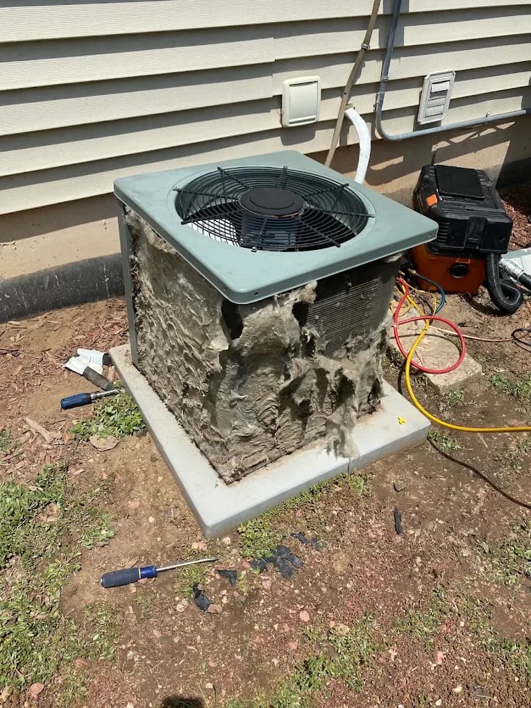 The coil of an outdoor air conditioning condenser packed with matted lint and debris before a maintenance clean, with the fan grille lifted clear of the cabinet